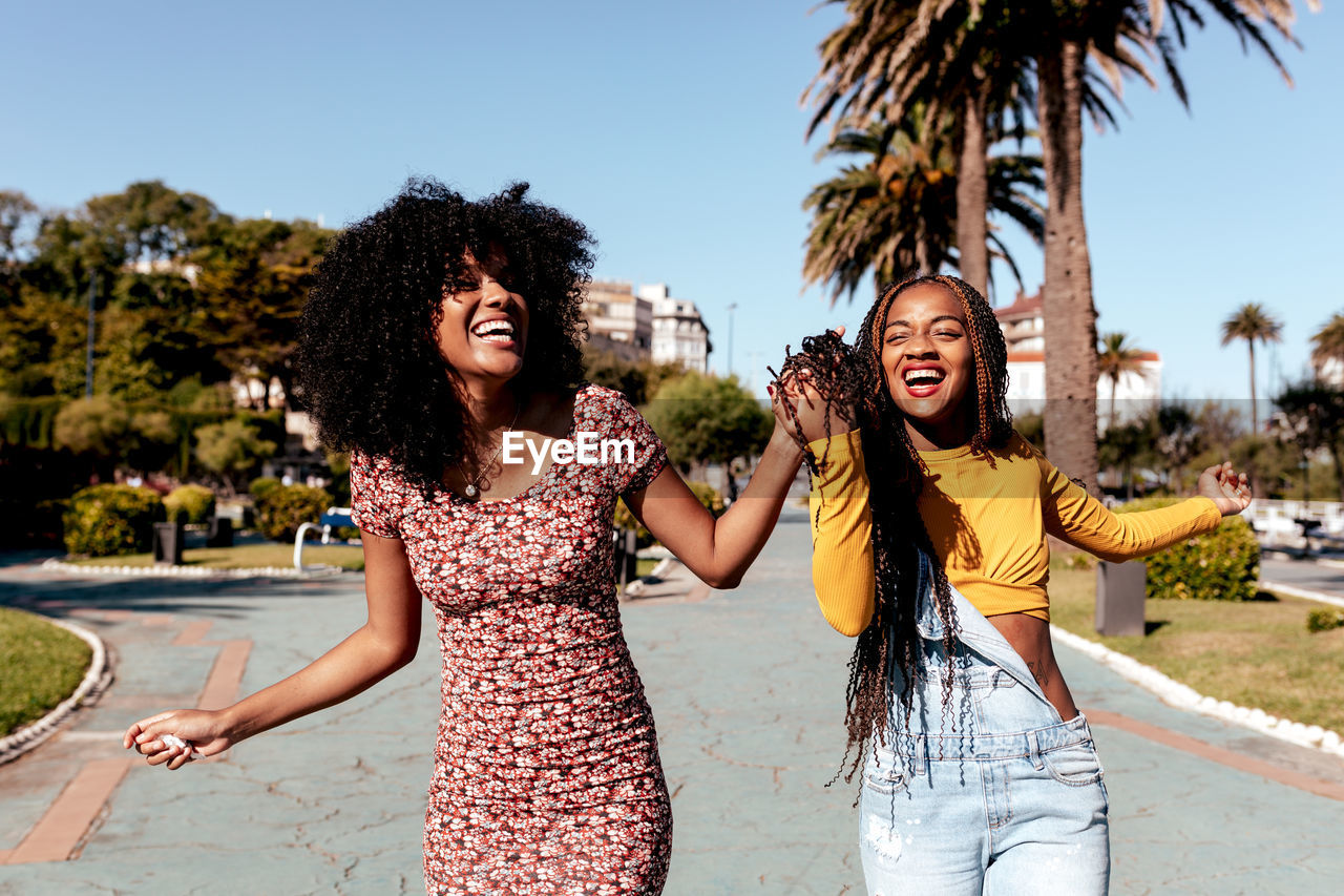 Delighted black woman with braids holding hand of african american female best friend with curly hair while walking along street in tropical city on sunny day