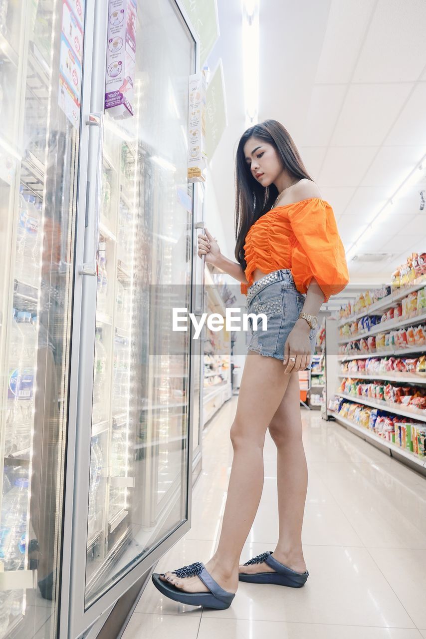 Side view full length of young woman opening shelf door in supermarket