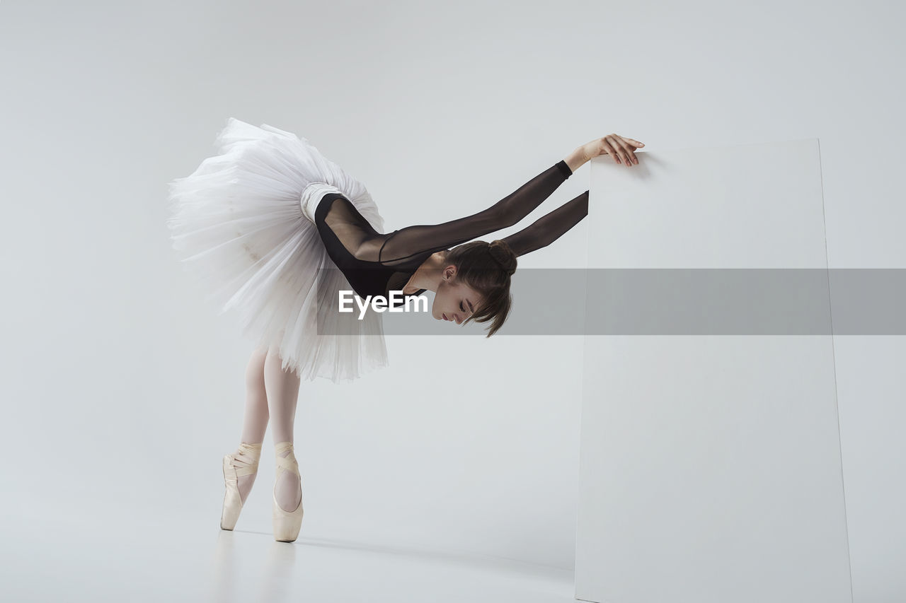 A ballerina in a black bodysuit and tutu standing on pointe shoes stretches leaning forward