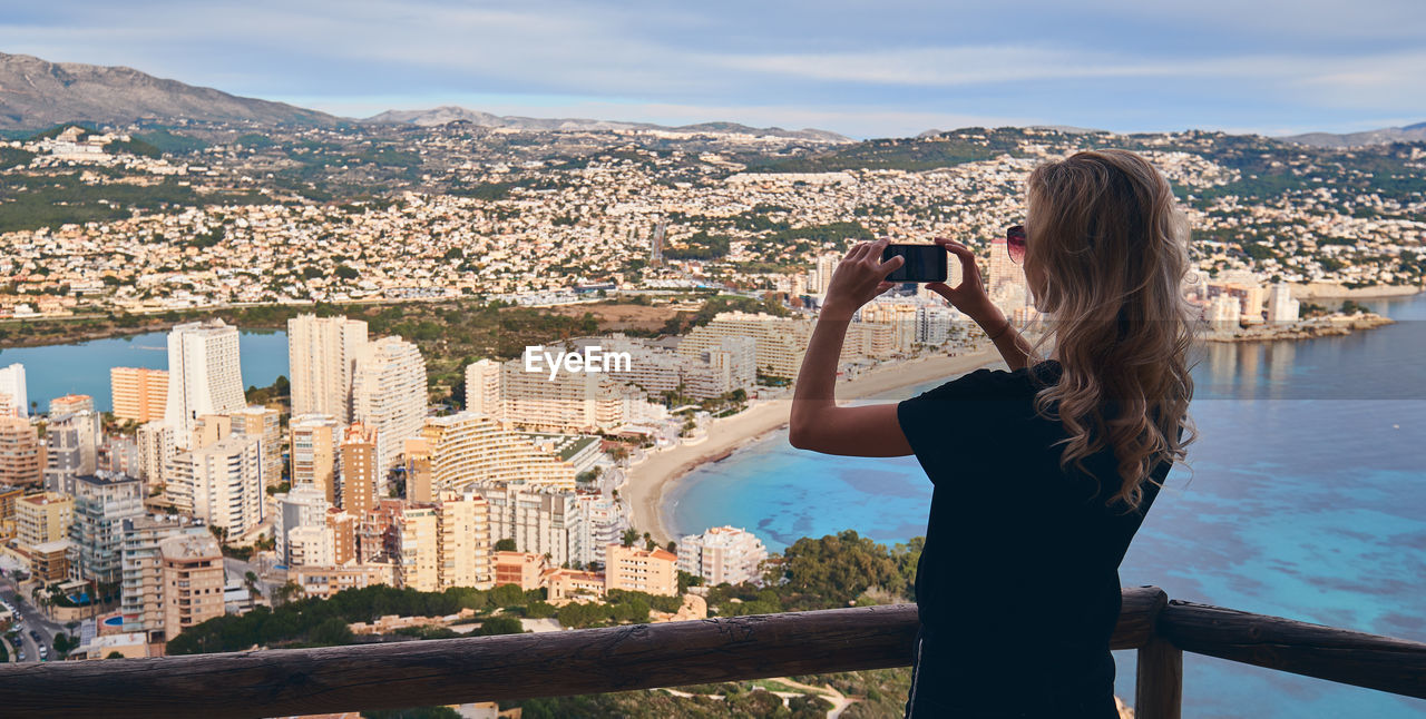 REAR VIEW OF WOMAN PHOTOGRAPHING THROUGH CITYSCAPE