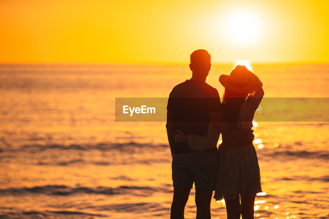 silhouette woman standing at beach against orange sky during sunset