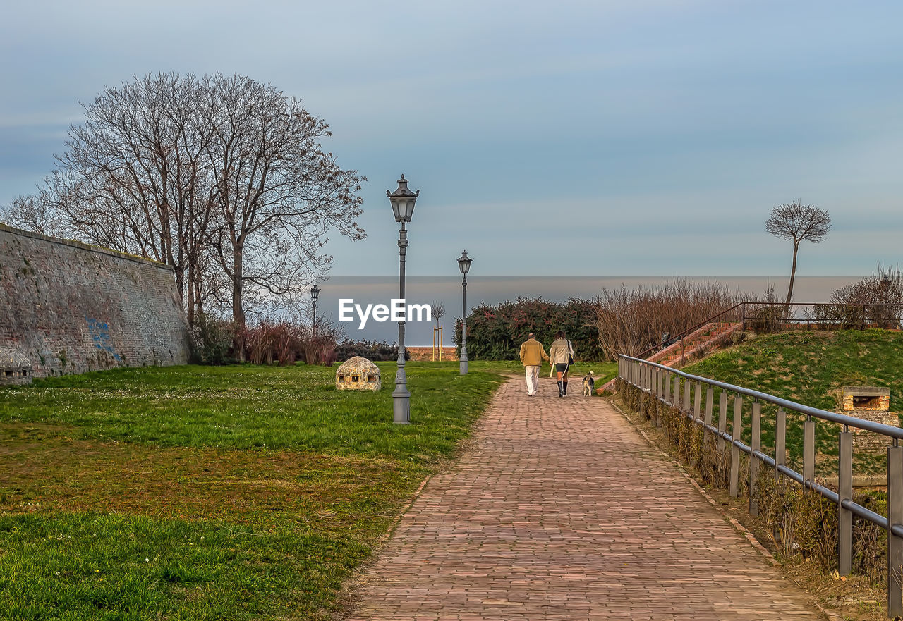 plant, sky, tree, rural area, nature, footpath, walkway, grass, the way forward, architecture, cloud, walking, day, outdoors, built structure, bare tree, landscape, field, waterway, land, women, men, morning, mammal, adult, group of people, beauty in nature, hill, animal themes, flower, lifestyles