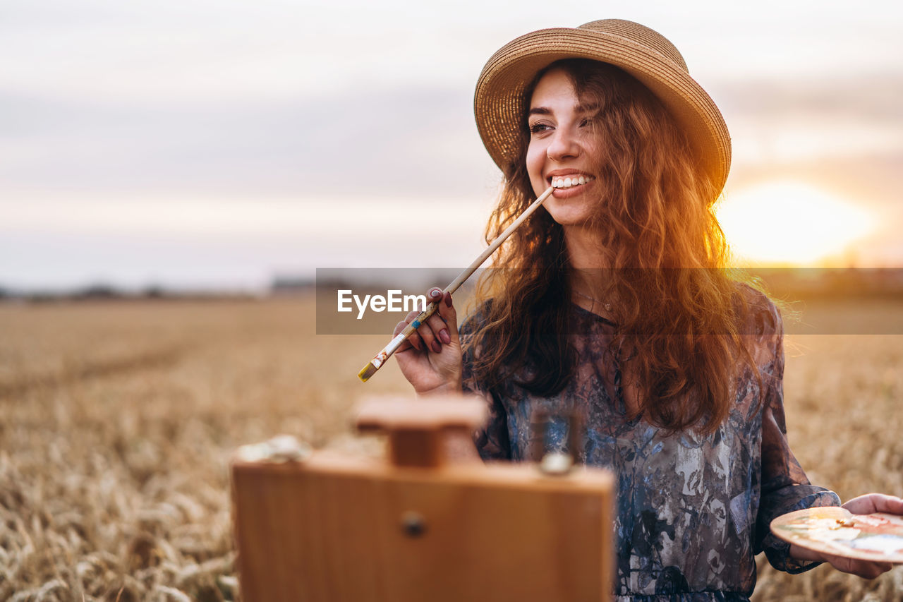 A young woman with curly hair and wearing a hat is painting in nature. 