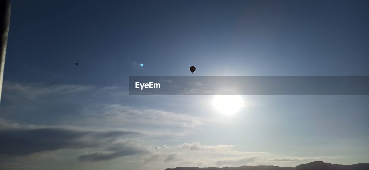 sky, flying, mid-air, nature, cloud, beauty in nature, hot air balloon, scenics - nature, mountain, balloon, horizon, adventure, sunlight, air vehicle, sun, environment, tranquility, astronomical object, tranquil scene, blue, outdoors, sports, silhouette, transportation, low angle view, extreme sports, no people, dawn, mountain range, day, landscape, sunbeam, travel, sunrise, moon, leisure activity, morning
