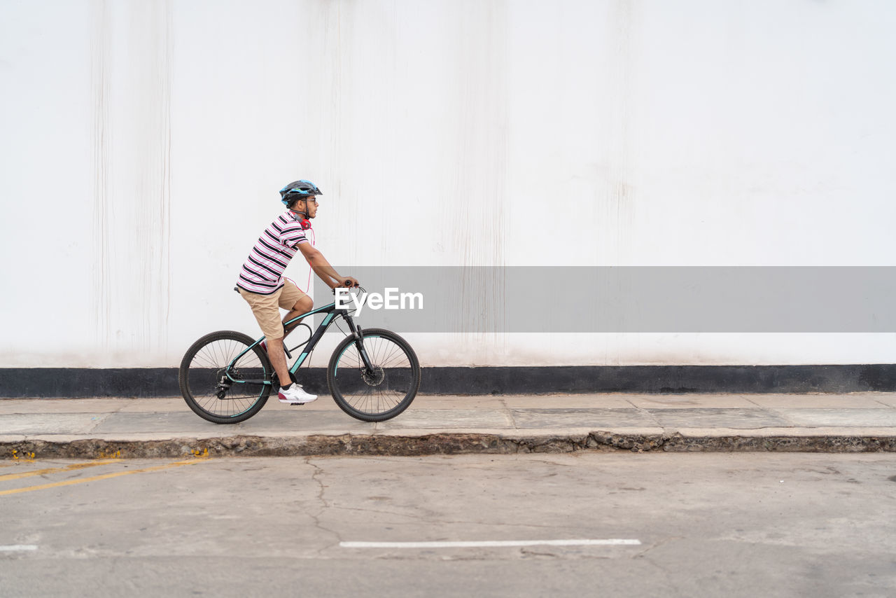 Full body side view of active male in protective helmet riding bicycle on sidewalk along road on street of city