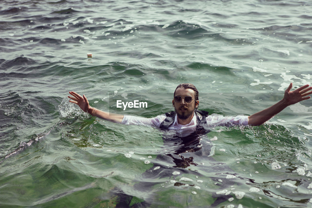 Man with a beard and sunglasses in clothes a vest and a white shirt swims in the sea among the rock