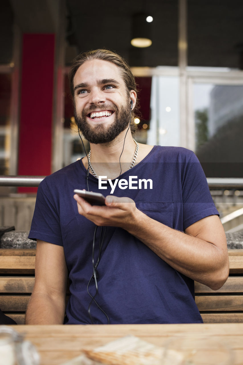 Smiling man listening music through smart phone while sitting at sidewalk cafe
