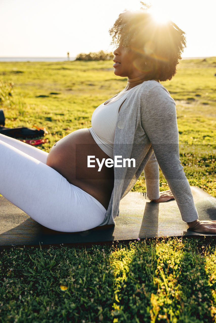 Side view of pregnant woman sitting on exercise mat over grass during training