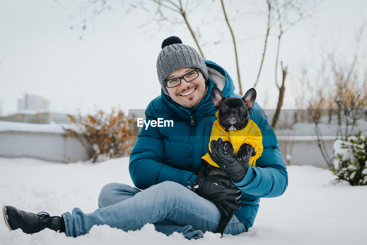 Portrait of man with dog sitting on snow covered field