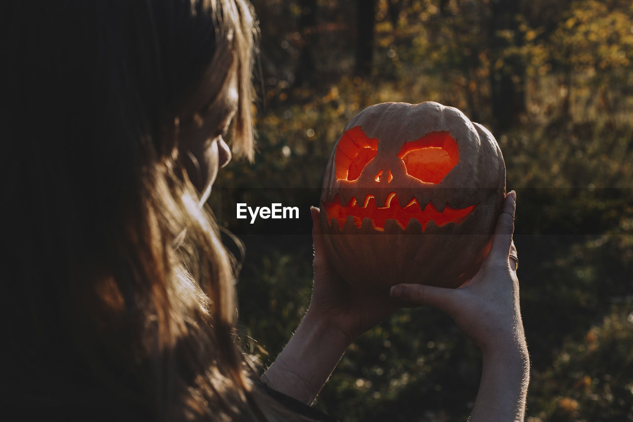 Young woman looking at jack o' lantern while standing in forest during halloween