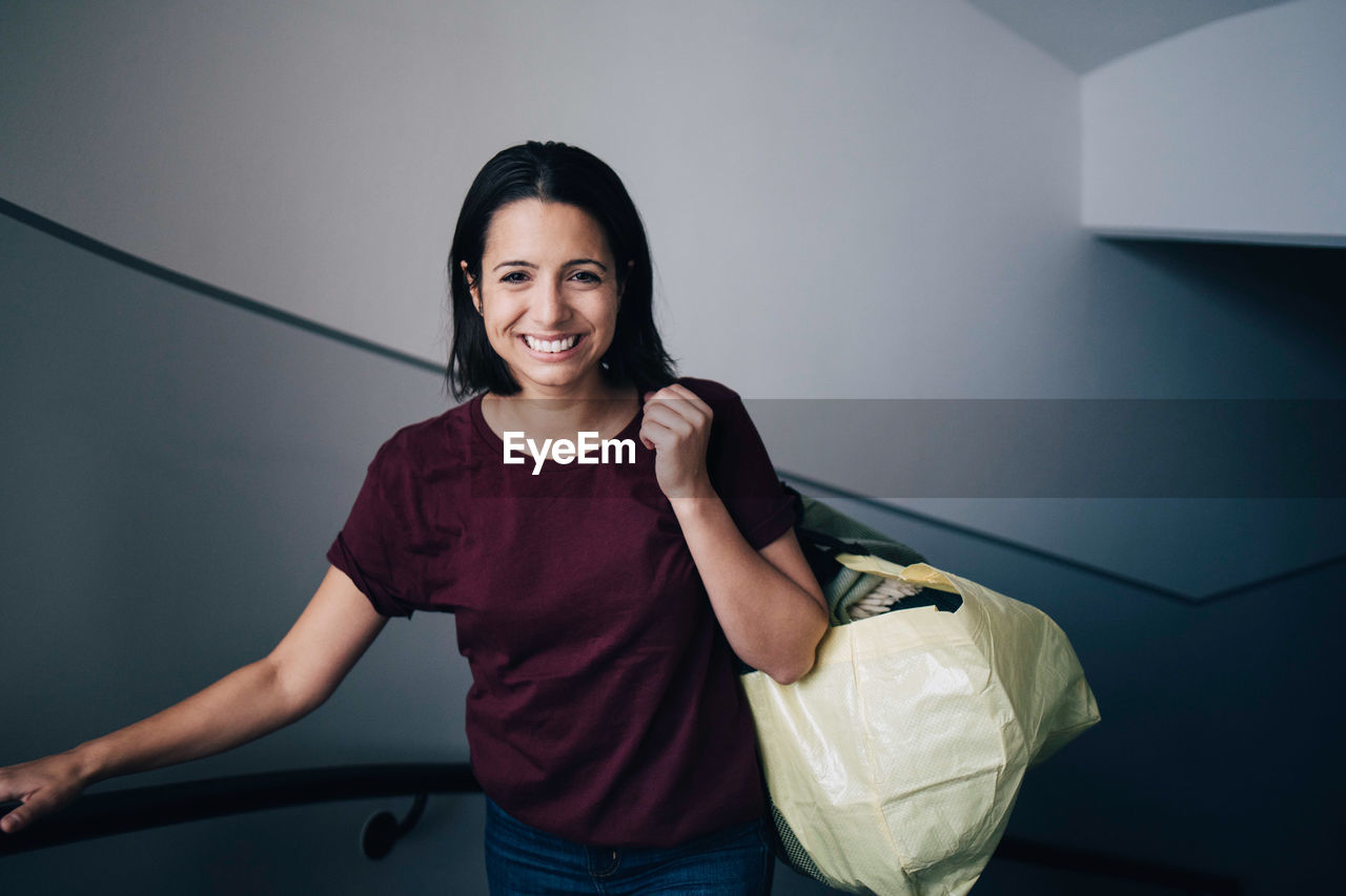 Portrait of smiling woman carrying bag while standing by wall