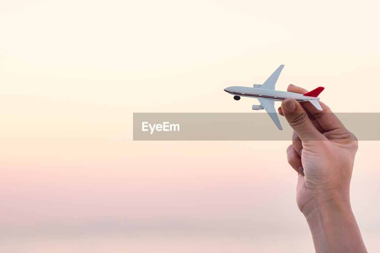 Cropped hand holding toy airplane against sky during sunset