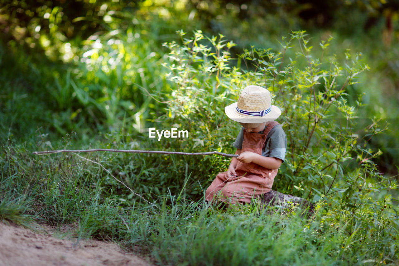 Cute european boy in hat and overalls in summer with stick fishing rod fishes in the grass near 