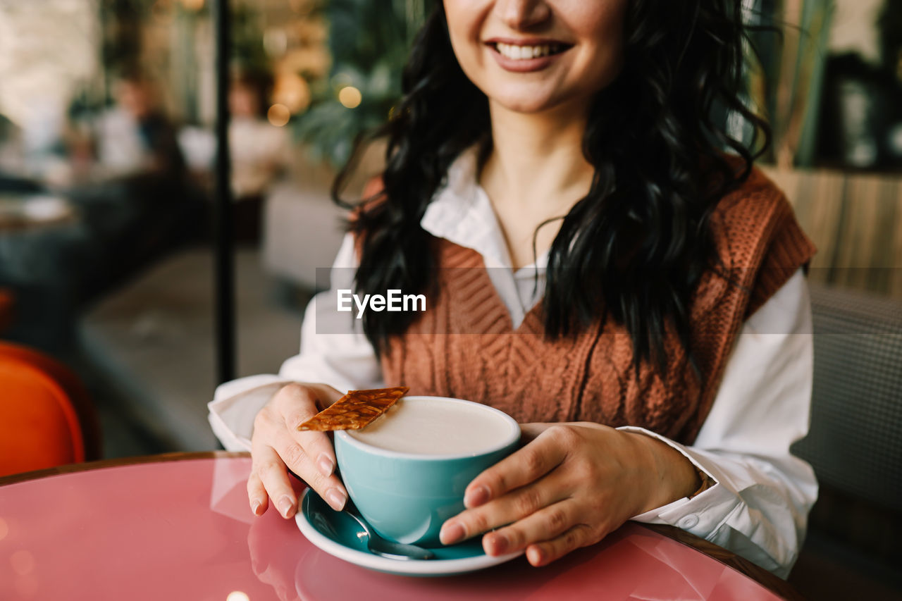 A young smiling woman enjoys a coffee drink while holding a mug while sitting in a cafe