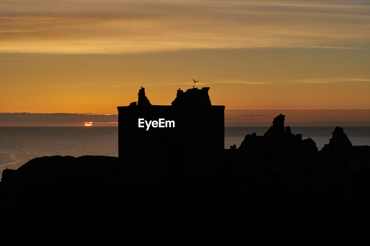 Silhouette buildings by sea against sky during sunset