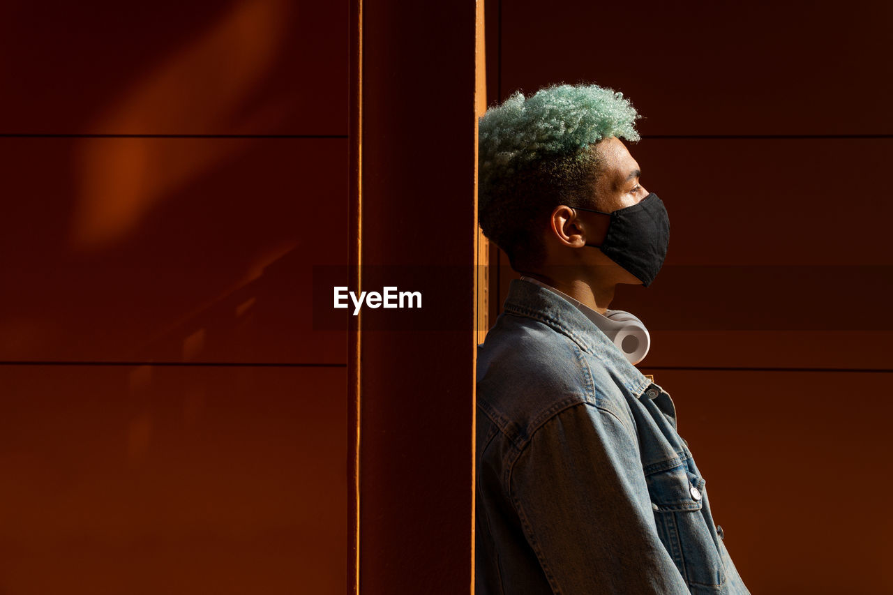 Side view of african american male hipster with blue curly hair wearing protective mask leaning on wall in city during coronavirus pandemic and looking away