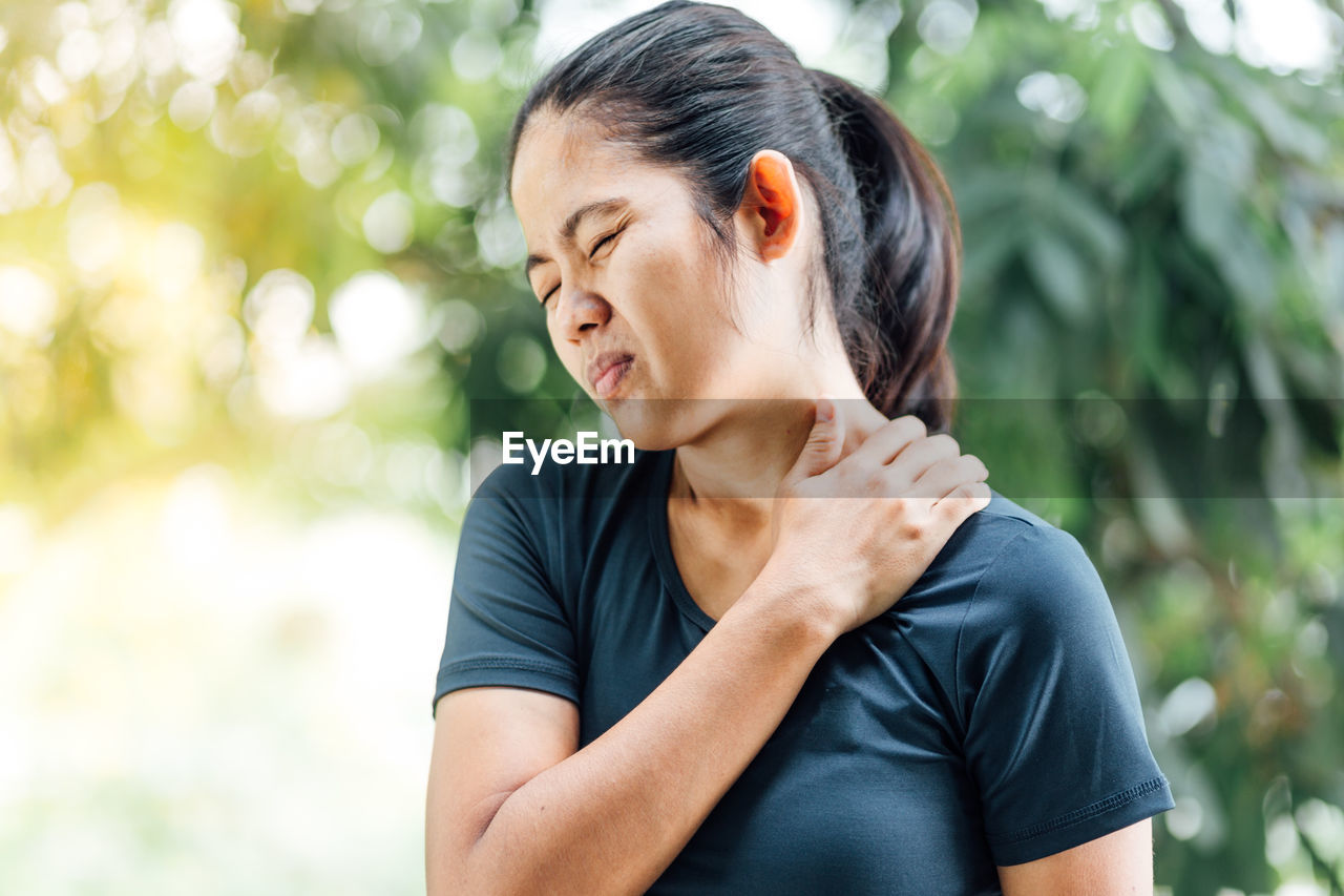Woman with eyes closed touching shoulder in pain against trees