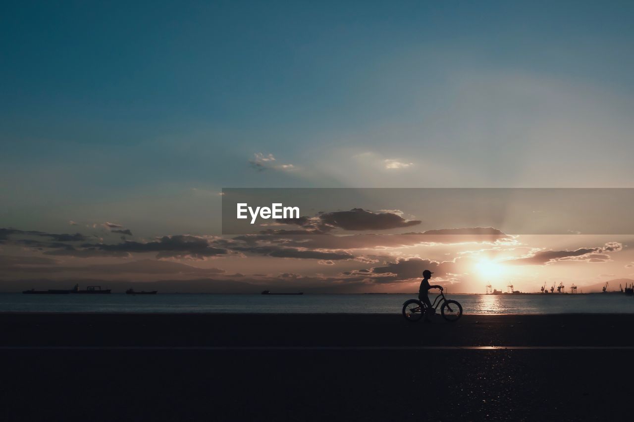 Man riding bicycle by sea against sky during sunset