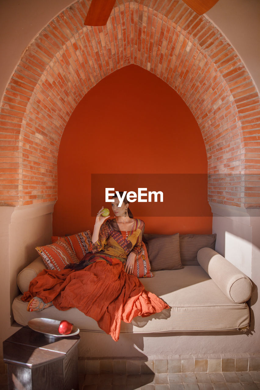 Full length of woman sitting on sofa against orange wall