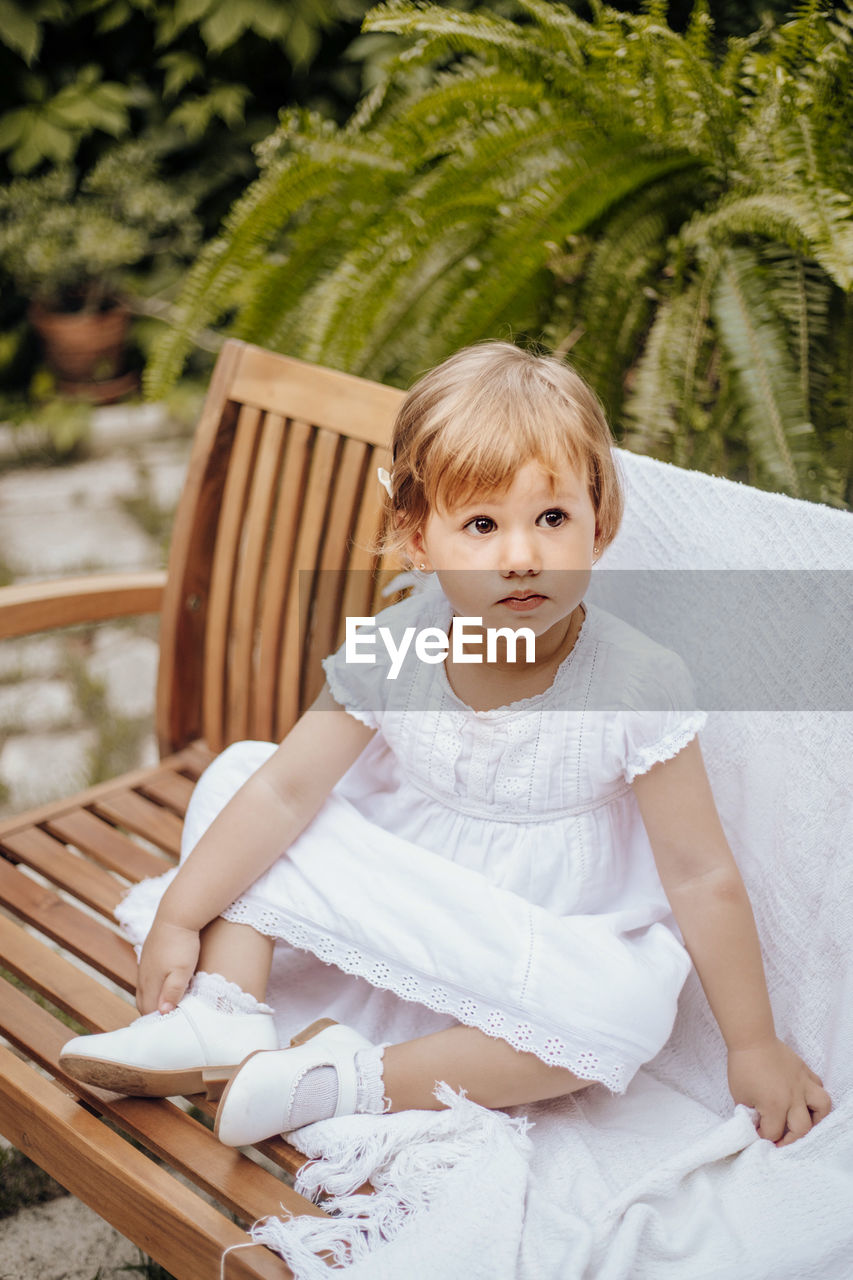 Little blonde girl sits on a wooden bench in the garden. child is wearing a dress. childhood