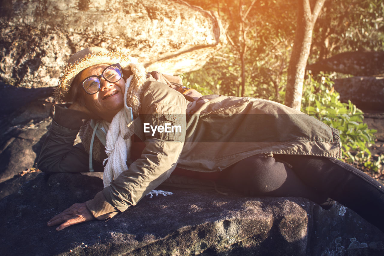 Portrait of smiling senior woman lying on rock at forest