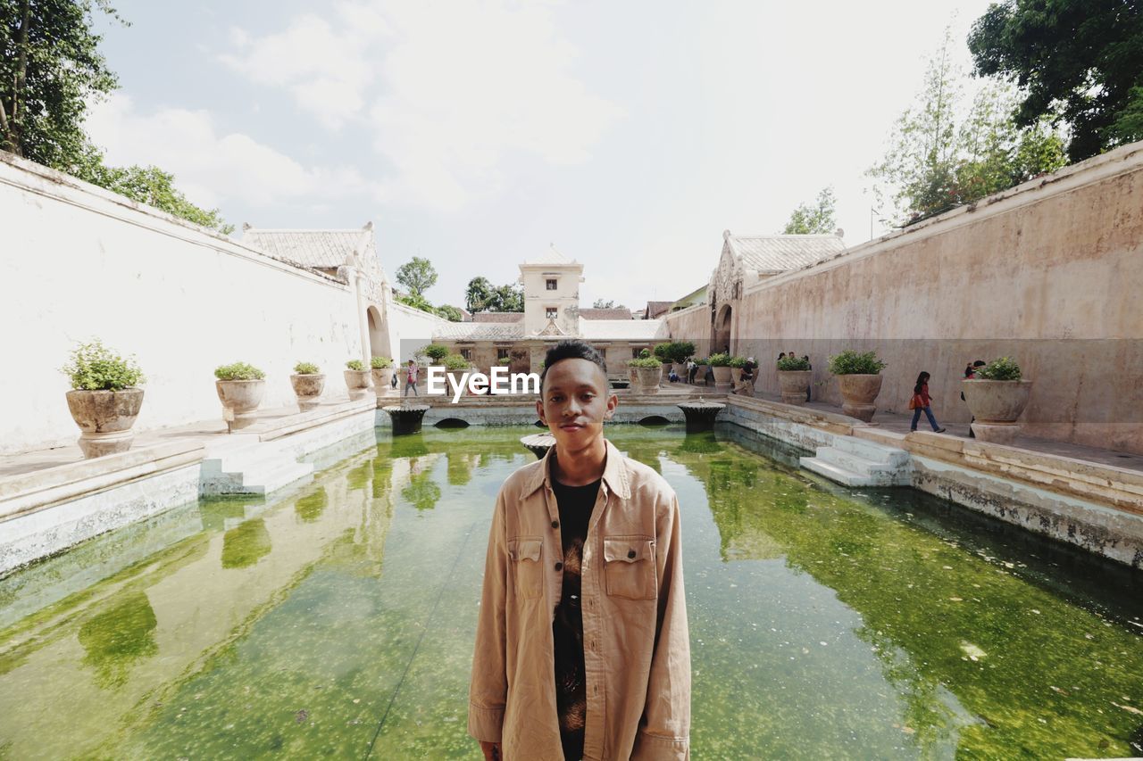 Portrait of young man standing by swimming pool against sky