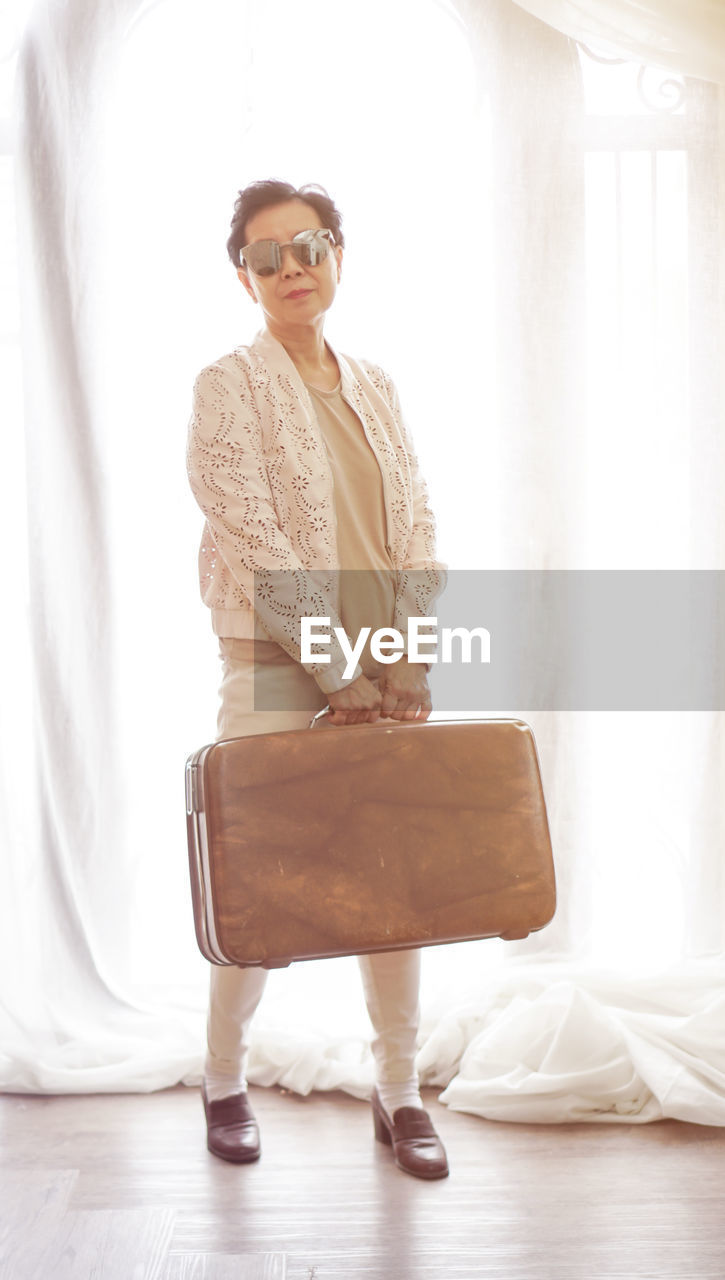 Portrait of woman holding suitcase while standing against window at home