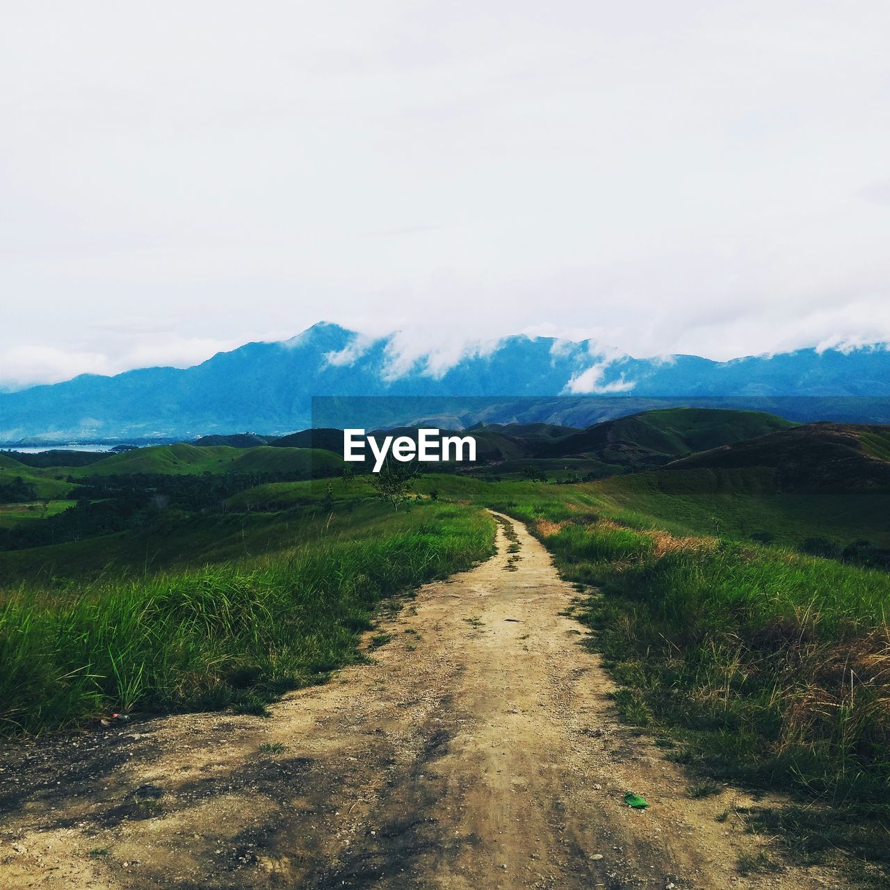 Dirt road amidst grassy field leading towards mountains against cloudy sky