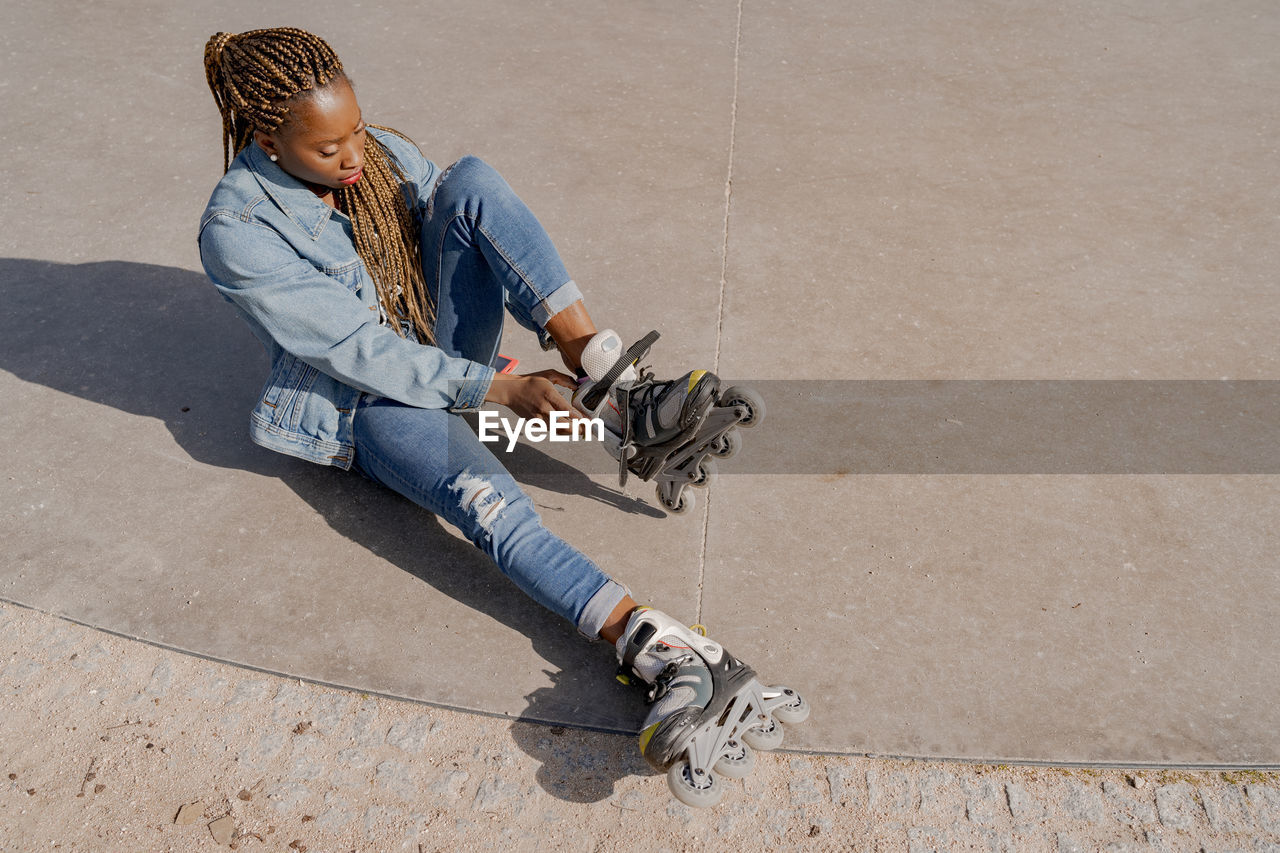 From above of african american female in denim outfit putting on rollerblades on sunny day in skate park