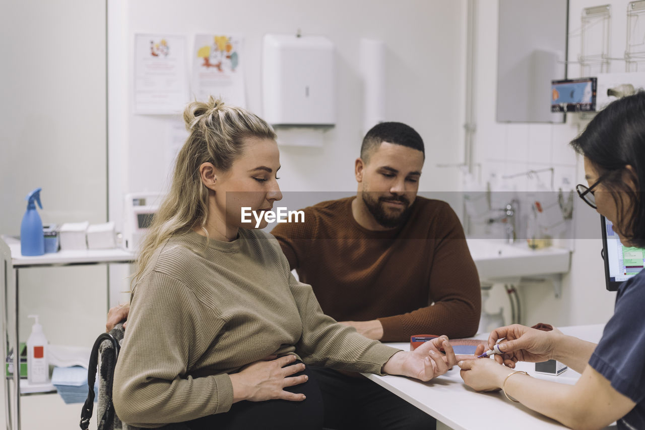 Gynecologist doing sugar test of pregnant woman sitting by man at desk in medical clinic