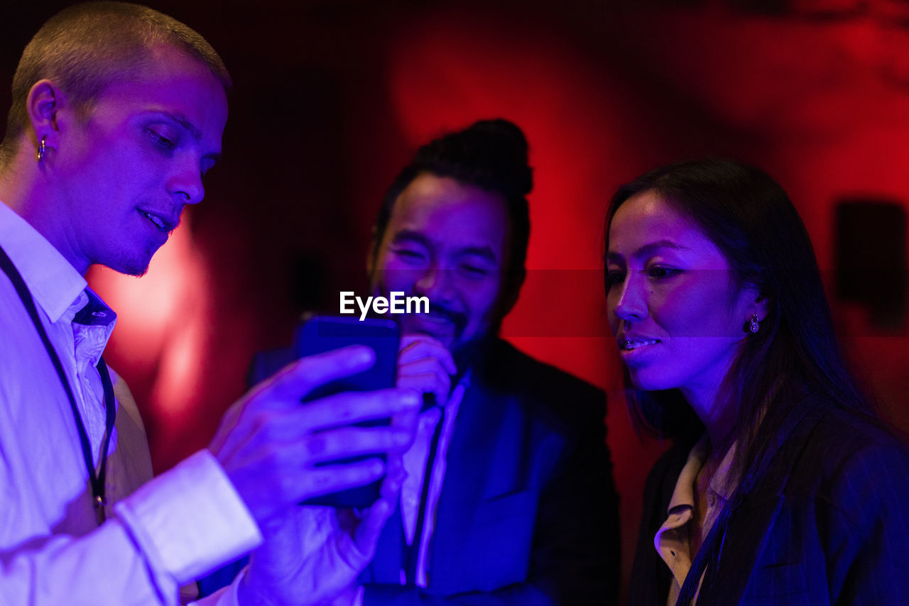 Businessman sharing mobile phone with male and female colleagues in illuminated workshop