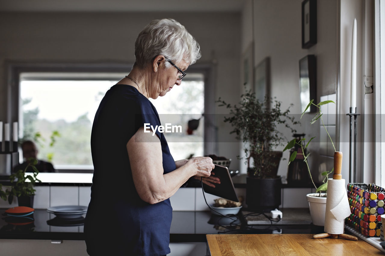 Side view of senior woman charging digital tablet in kitchen at home