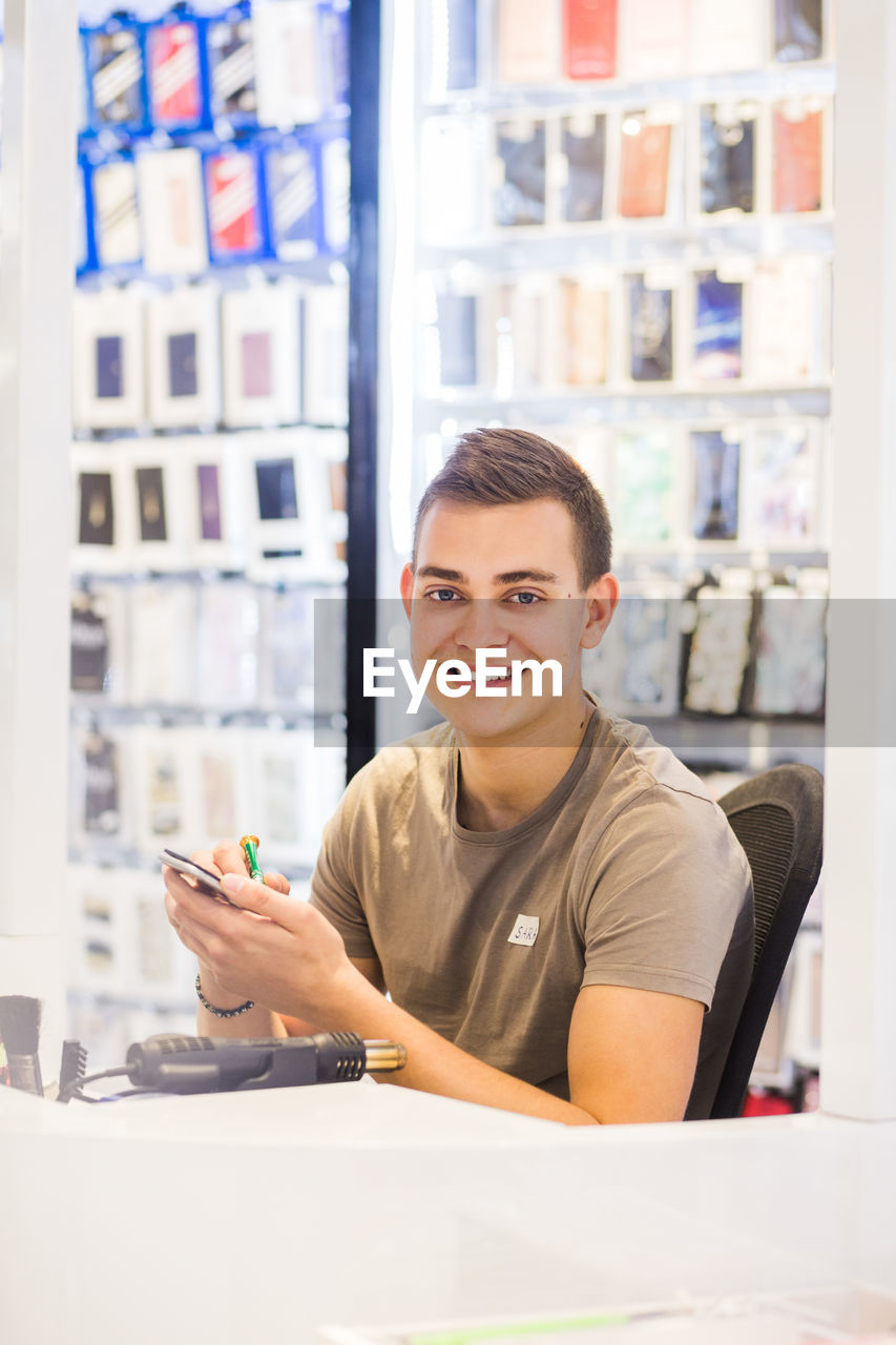 Portrait of smiling young male trainee repairing mobile phone while sitting at illuminated desk in store