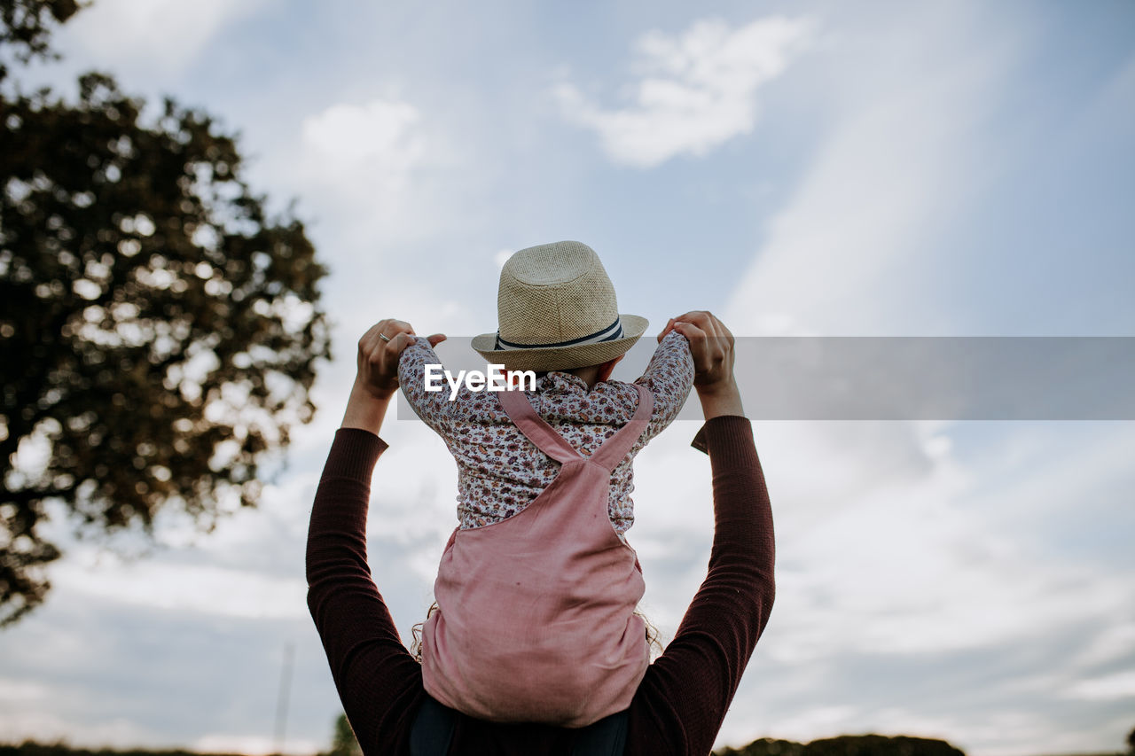 Mother carrying daughter on shoulders against cloudy sky