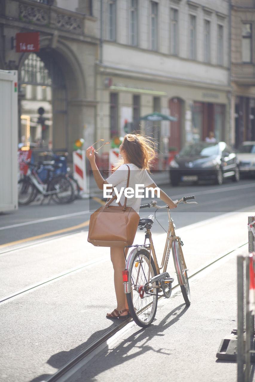 Side view of young woman holding sunglasses with bicycle on city street