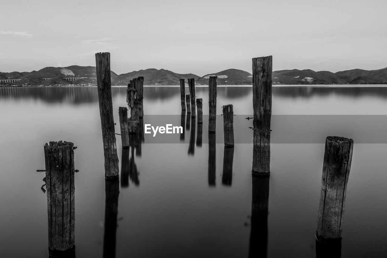 water, reflection, black and white, wooden post, post, monochrome, monochrome photography, wood, sky, tranquility, nature, no people, black, lake, tranquil scene, scenics - nature, darkness, beauty in nature, fog, white, outdoors, pier, architecture, horizon, environment, cloud, in a row, day, built structure, landscape