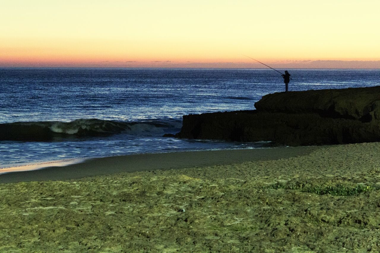 Person fishing while standing on cliff at beach against clear sky during sunset