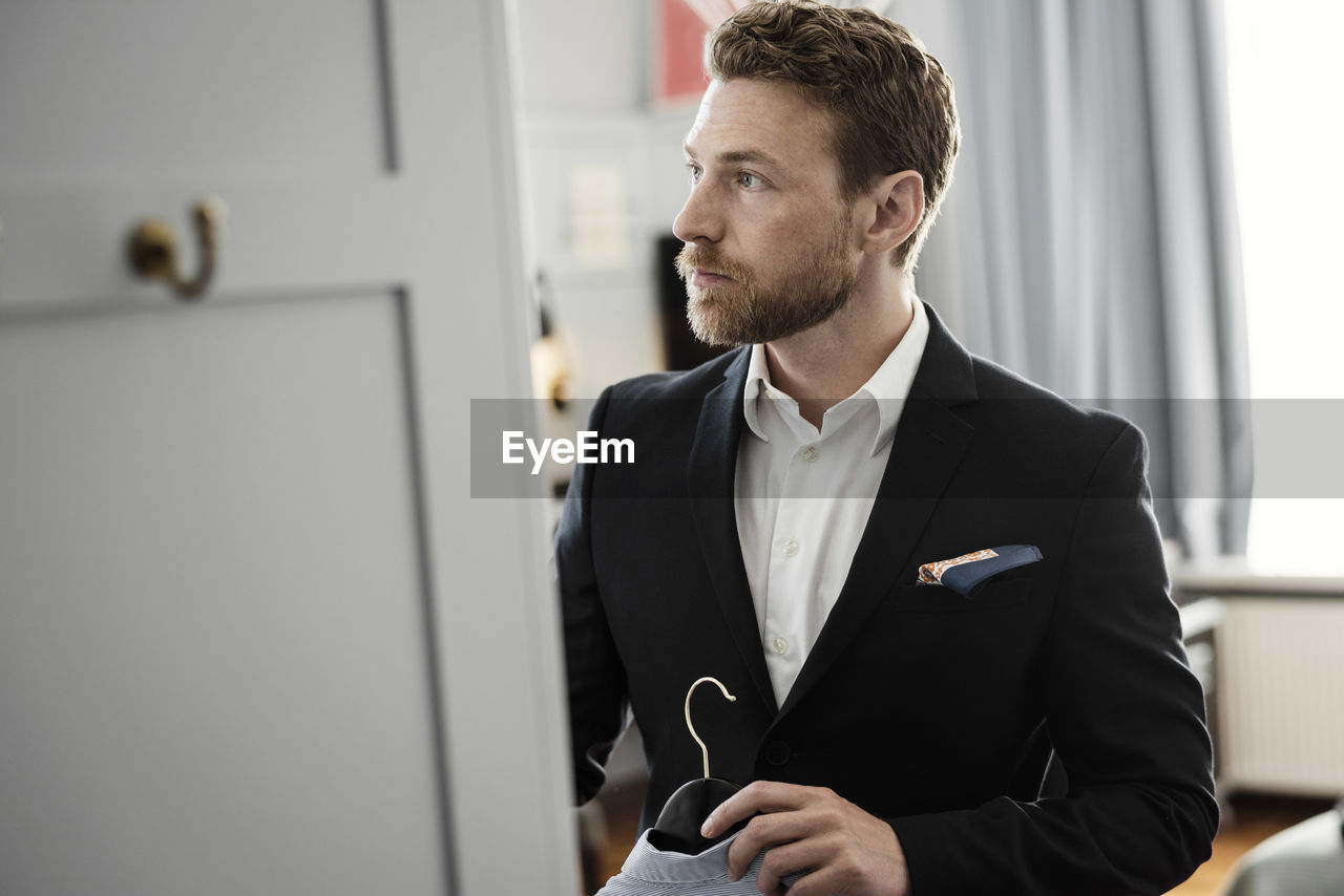 Mature businessman holding coathanger standing in hotel room