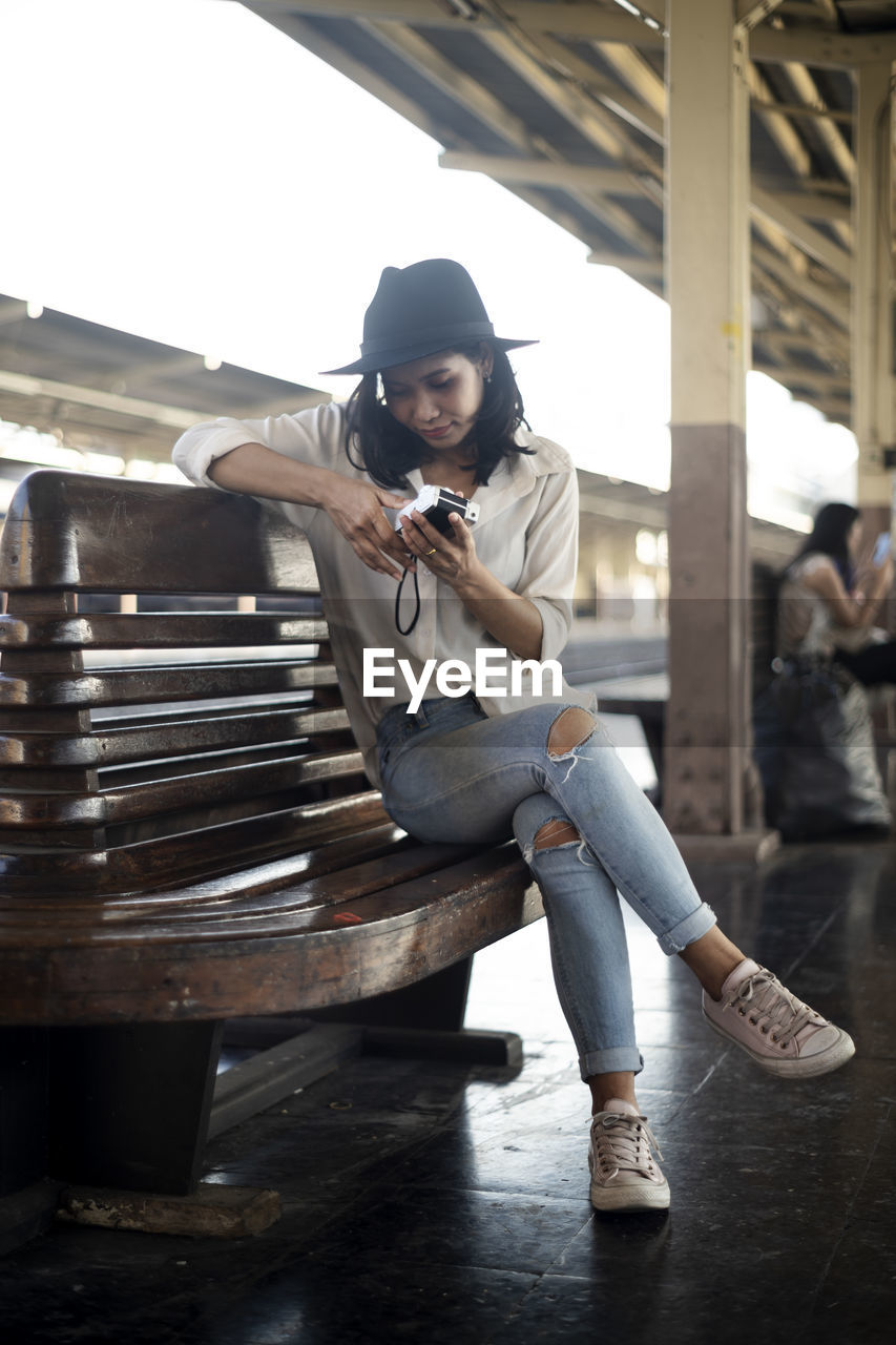 Full length of woman holding camera sitting at railroad station platform