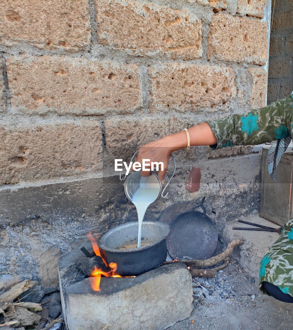 one person, fire, burning, flame, day, heat, nature, kitchen utensil, wall - building feature, wall, lifestyles, hand, outdoors, food and drink, container, holding, soil, iron, brick, food, adult, household equipment, architecture, women