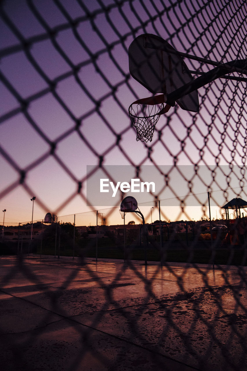 Silhouette basketball hoop seen through chainlink fence against sky during sunset