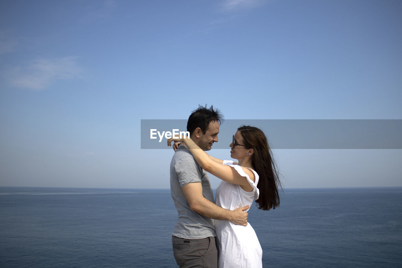 Young couple looking at each other standing in front of sea against sky