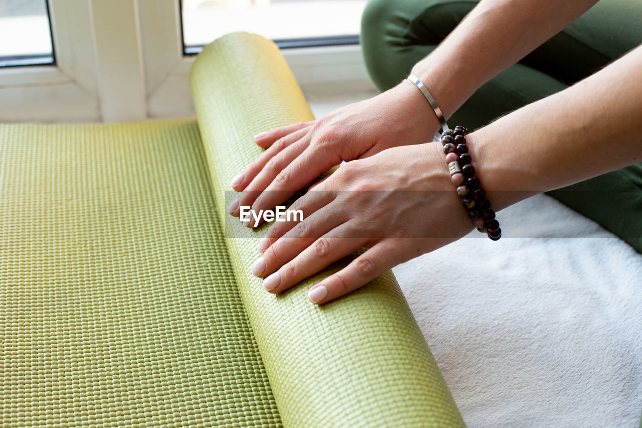 A hands slender woman twirls green rubber mat for fitness or yoga sitting  by the window.