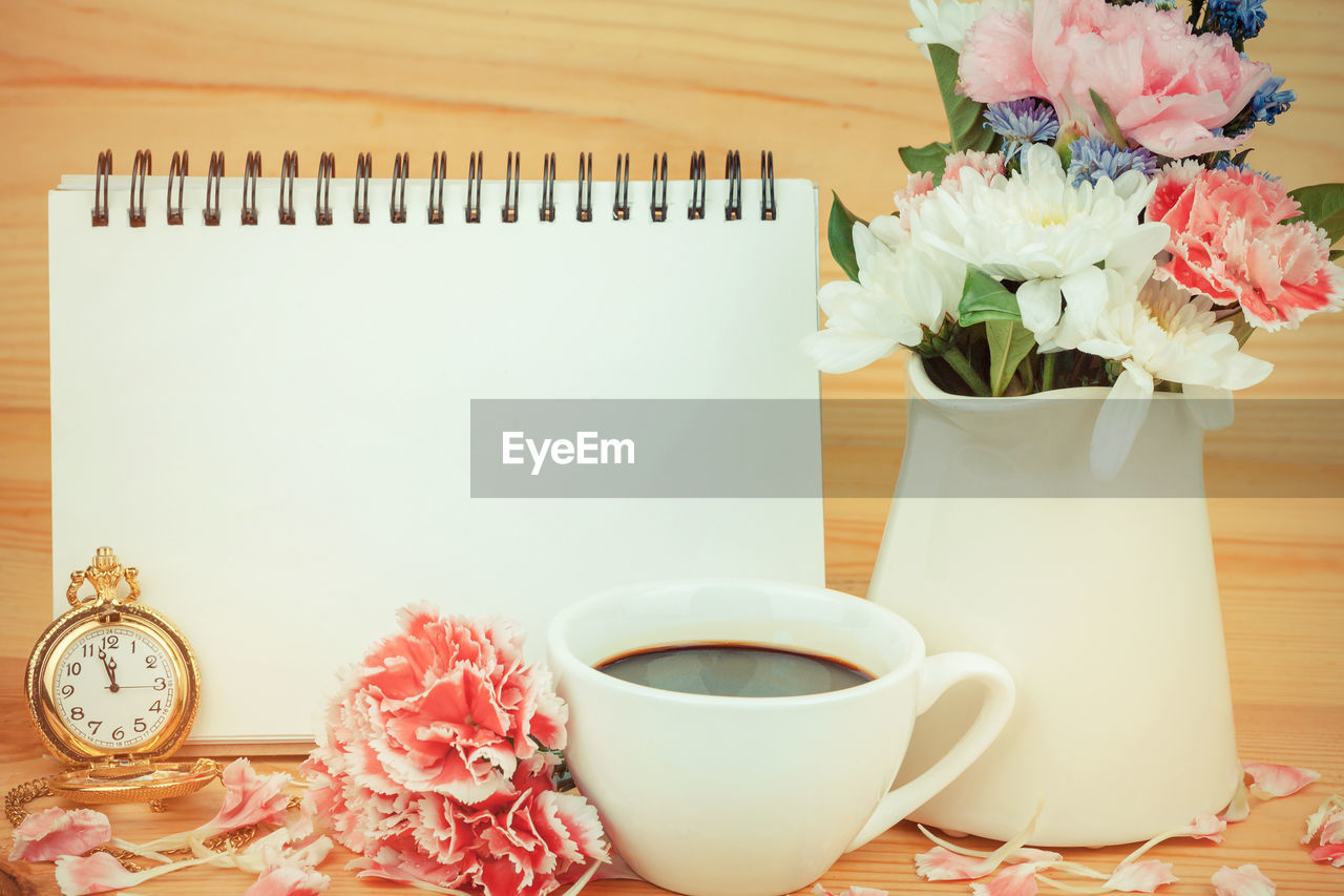 Close-up of coffee cup with flower vase and spiral notebook against wooden wall