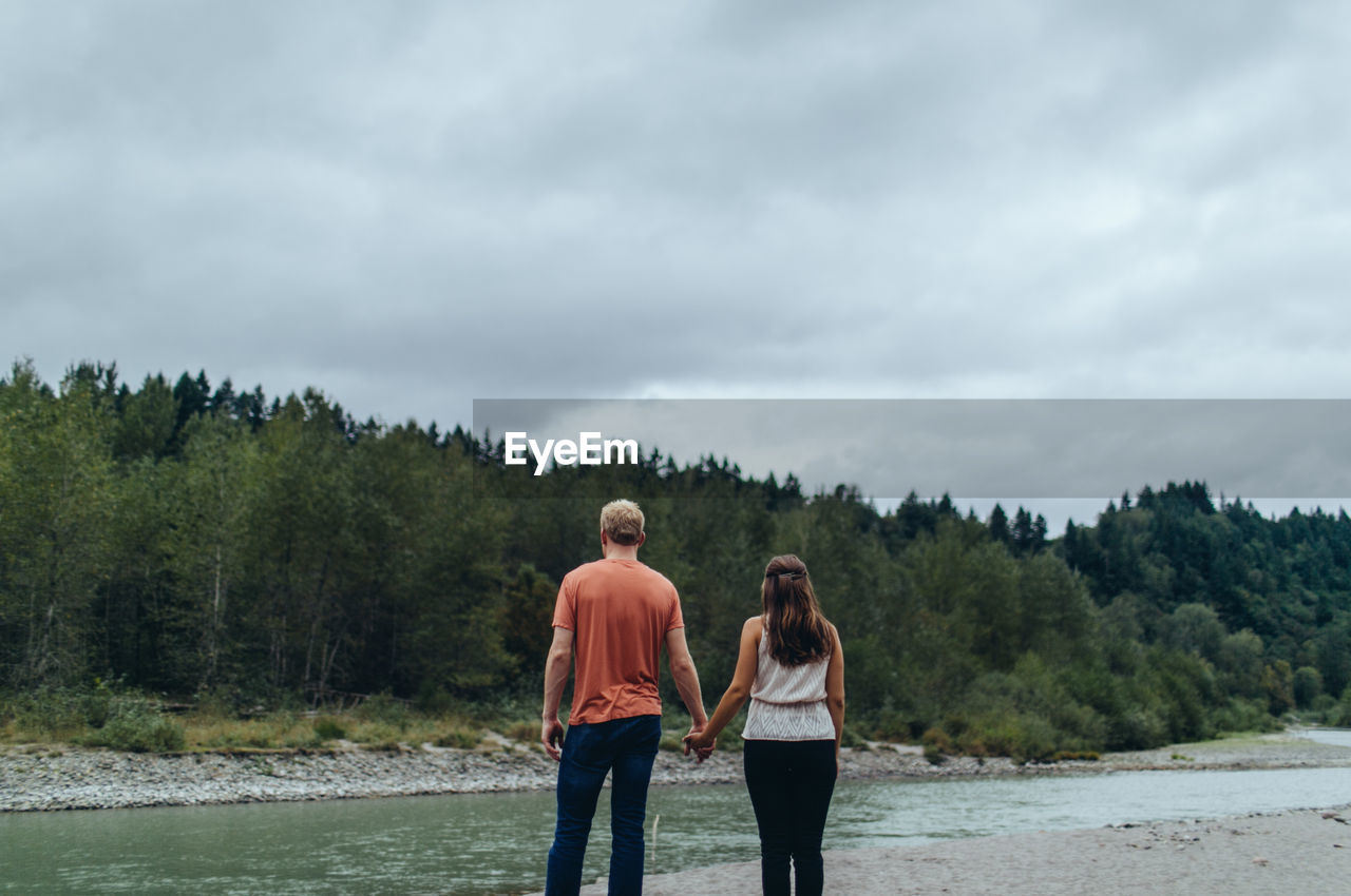 Rear view of couple standing by river in forest against cloudy sky