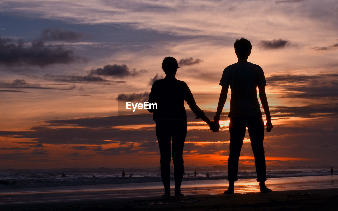 Silhouette of couple holding hands at beach against sky during sunset