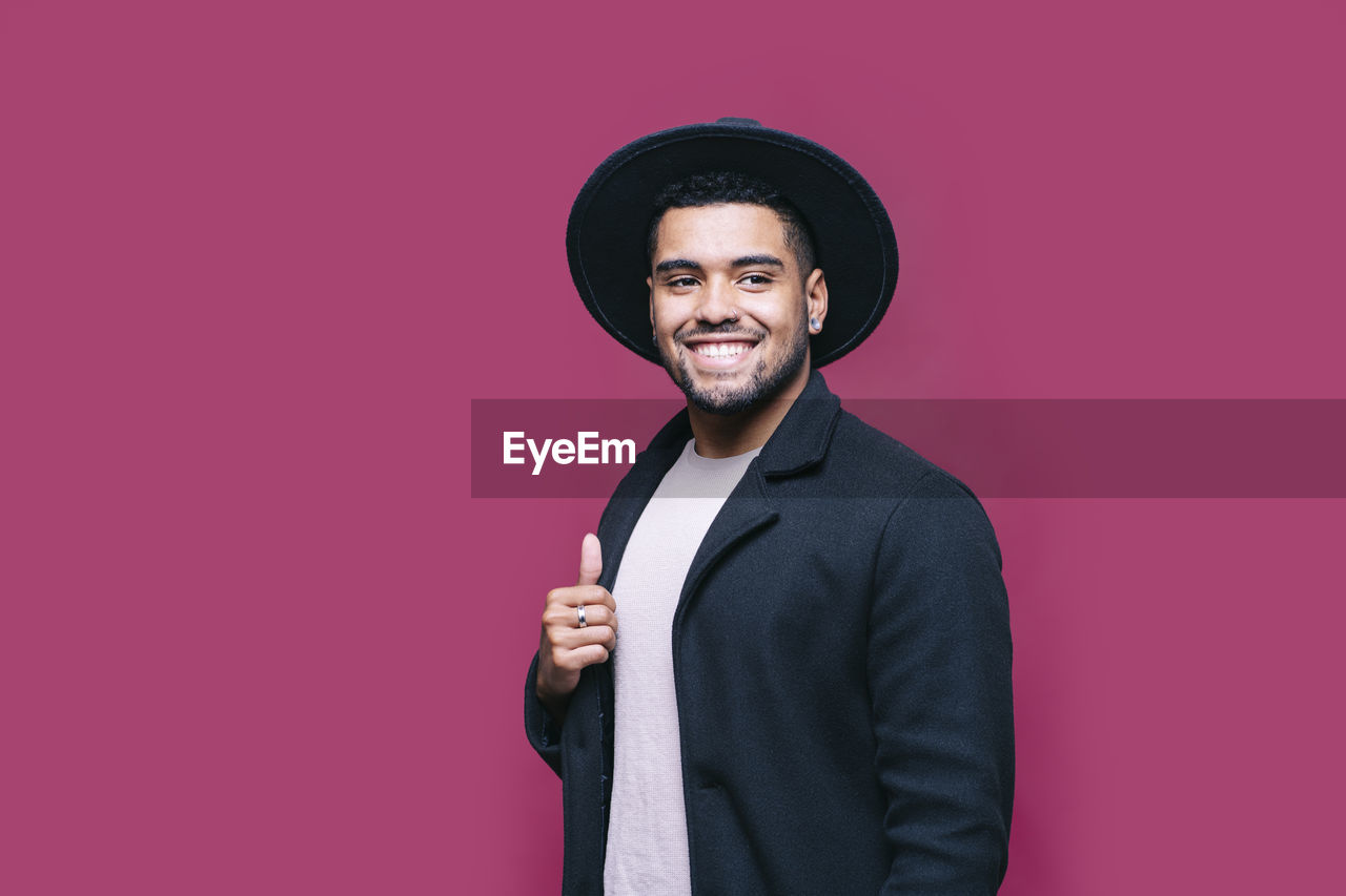 Smiling man in hat wearing blazer jacket while looking away against pink background