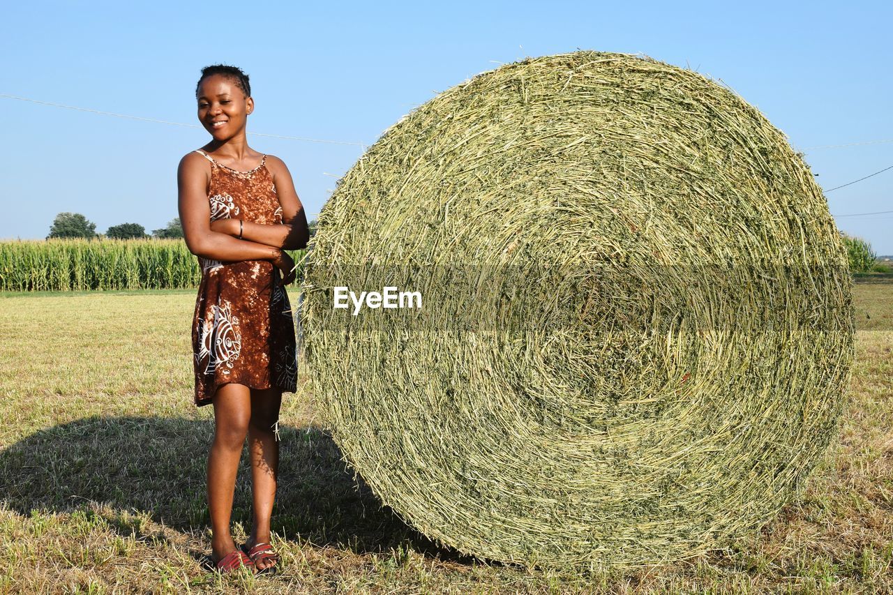 Full length of smiling woman standing by hay bale on field