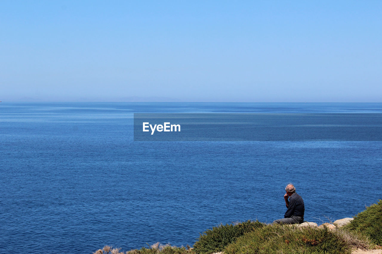 Rear view of man sitting on rock by sea against sky