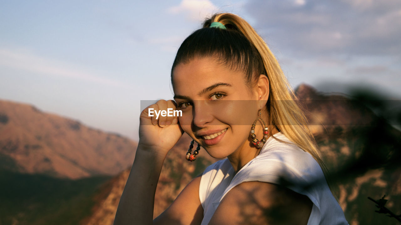 Portrait of smiling young woman sitting by rock formation against sky